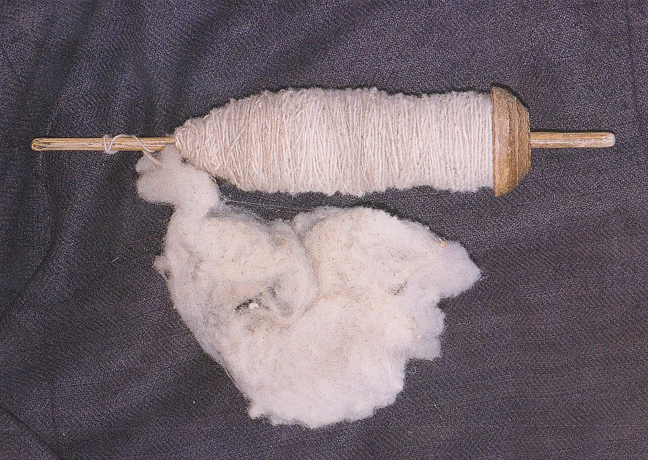 A wooden drop spindle with a whorl and hook hanging in mid-air with a length of handspun singles twisted yarn above it, against a light background