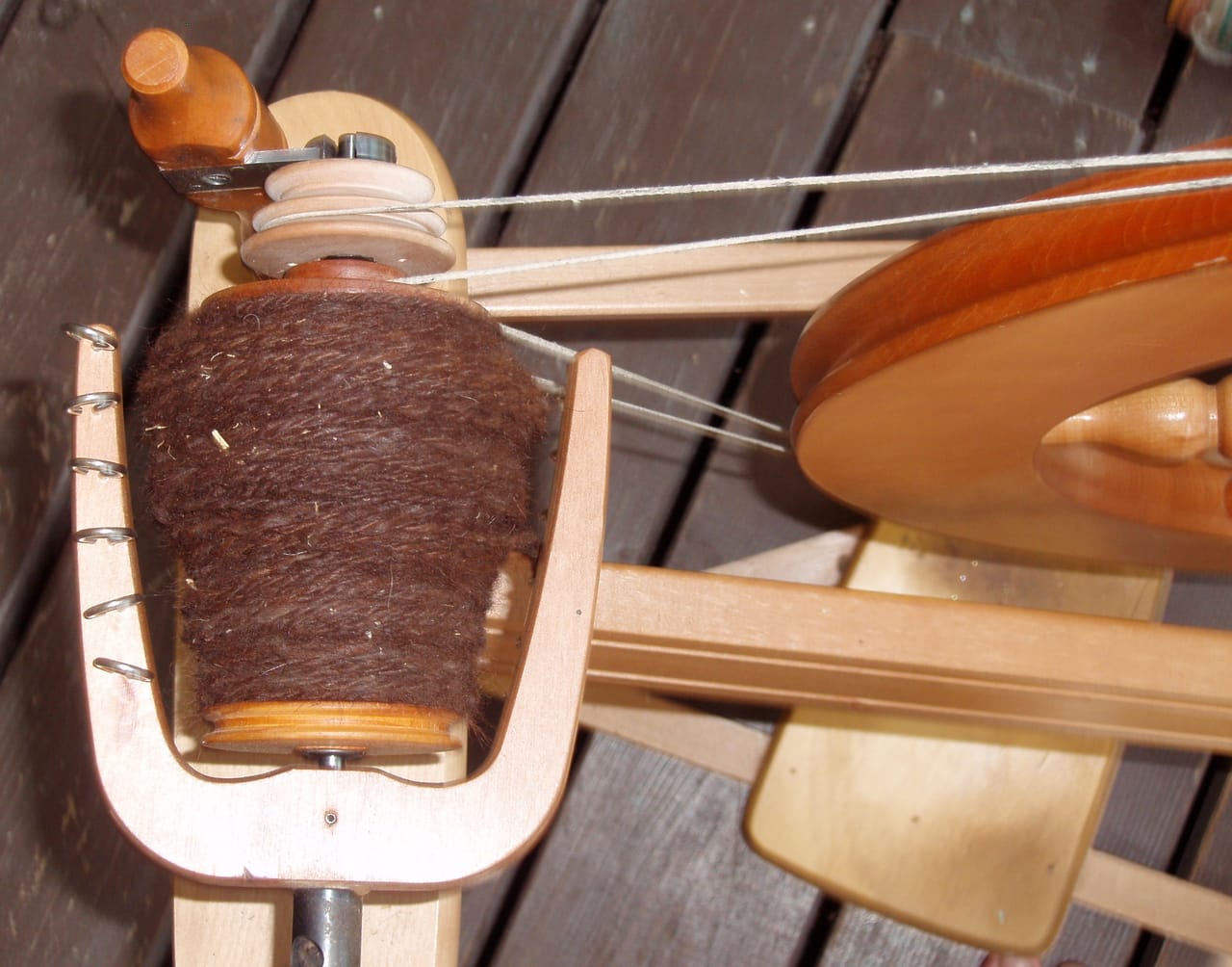 Close-up of a spinning wheel flyer-bobbin assembly showing the drive band around the flyer whorl, the bobbin seated on the spindle between the flyer arms, and the hooked flyer arms that guide yarn onto the bobbin