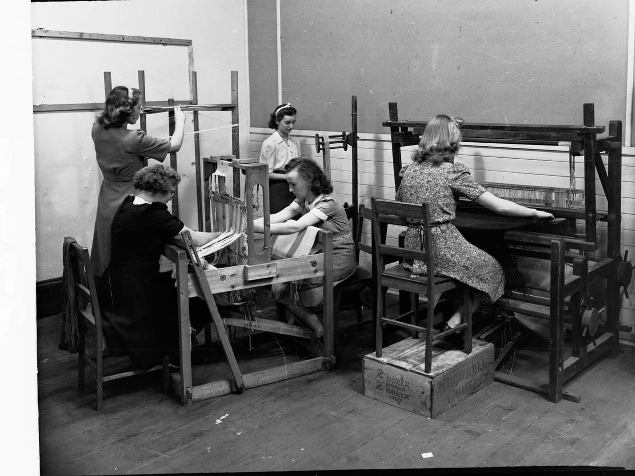 Young women weaving at looms in a school workshop, each working at a separate loom with colorful yarns strung as warp
