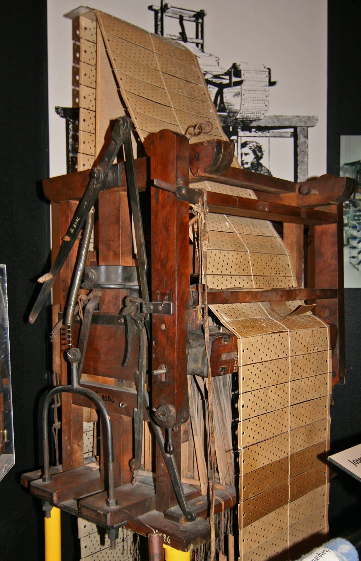 A historic Jacquard loom at a museum showing the punched-card pattern control mechanism mounted above the large weaving frame, illustrating automated shaft control