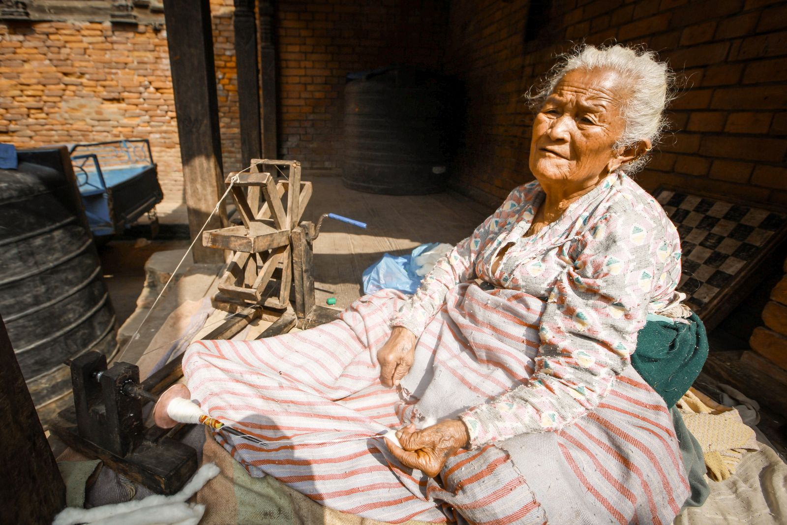 Elderly woman operating a traditional wooden treadle spinning wheel outdoors in a rustic setting