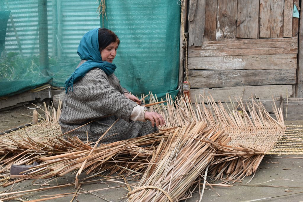 A woman weaving on a traditional loom, working the threads with practiced motions as cloth builds on the beam in a textured woven structure