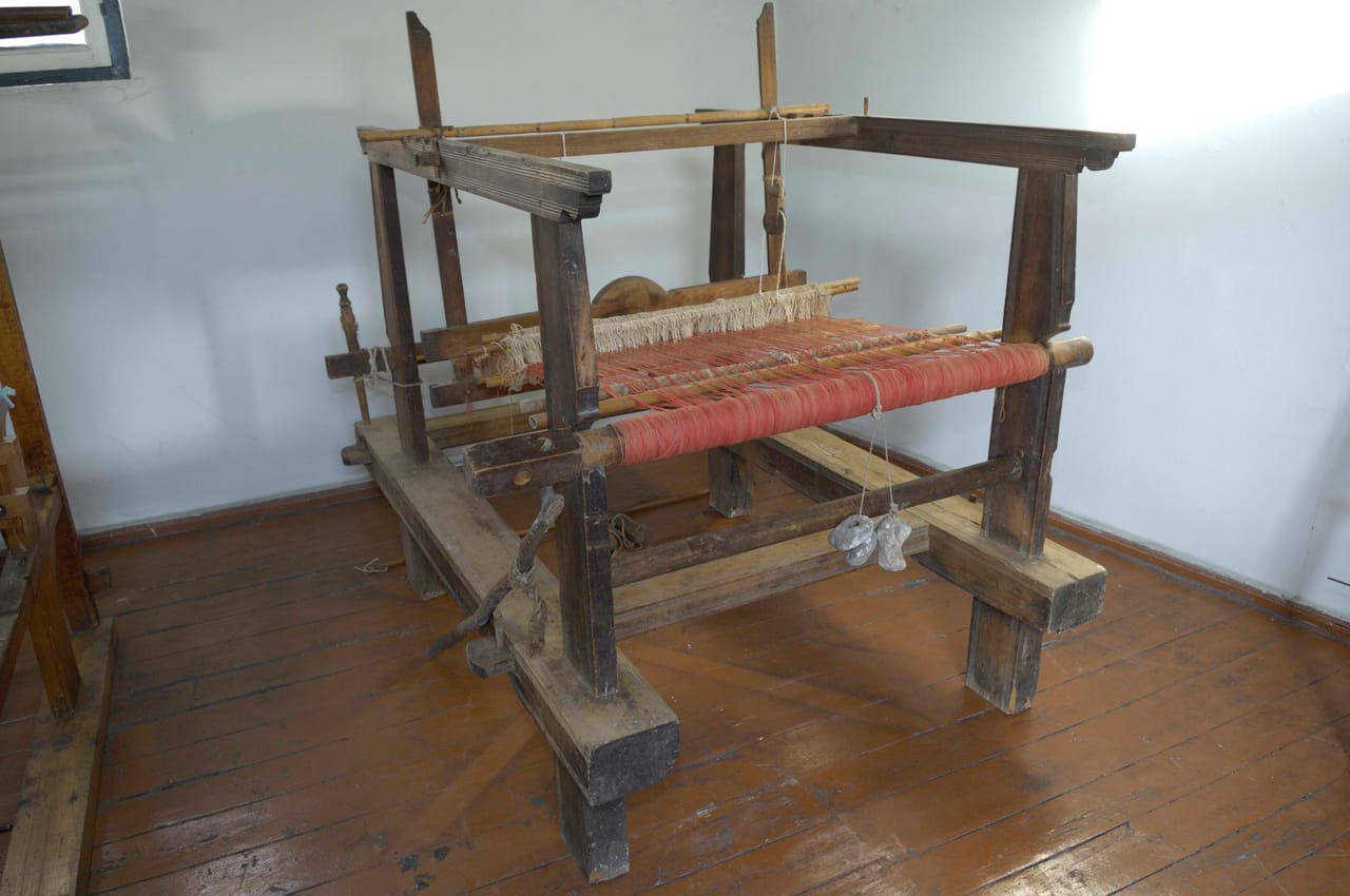 An 18th-century wooden floor loom from Crete with two harnesses, showing heddles suspended from the harness frames, warp threads running through, and the beater with its reed
