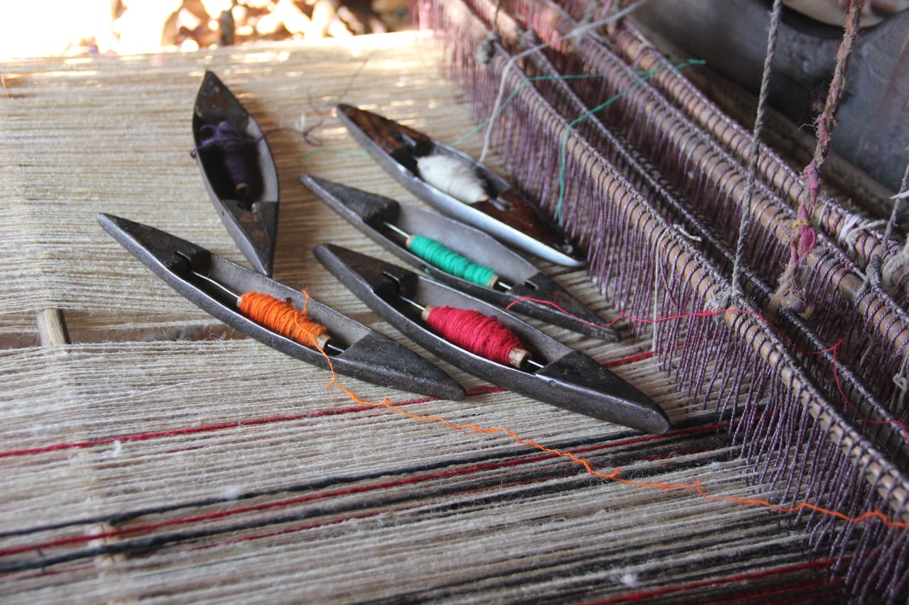 A wooden loom with a shuttle resting in the shed, showing the stick shuttle and weft yarn path through the open shed between raised and lowered warp threads