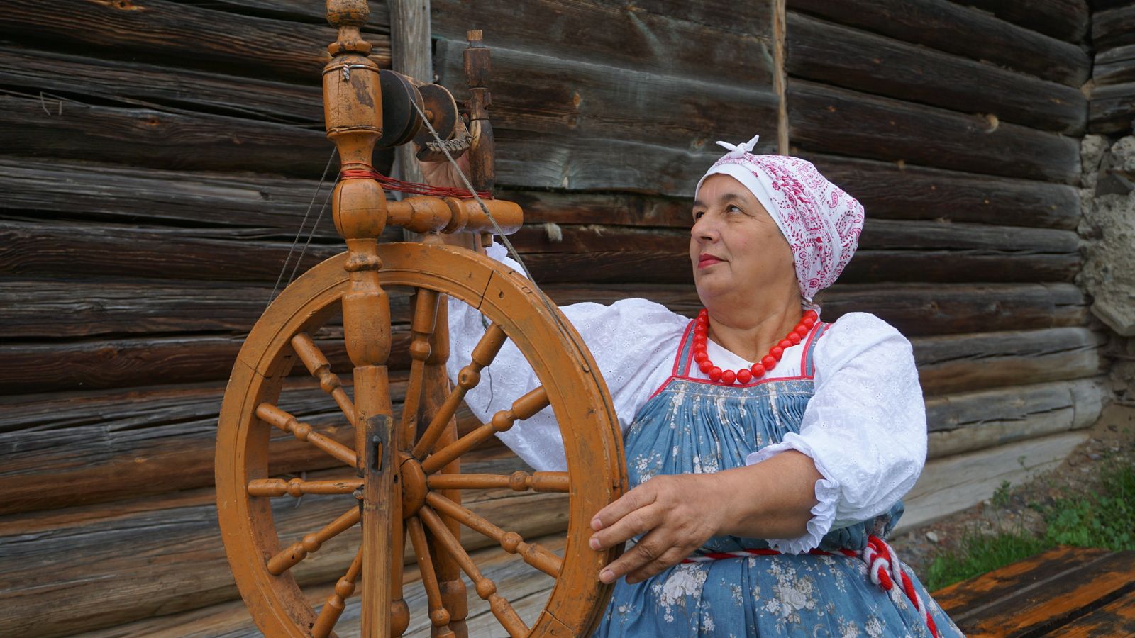 Elderly woman in traditional ornate dress spinning at a Saxony-style spinning wheel outdoors showing the long horizontal frame and large drive wheel
