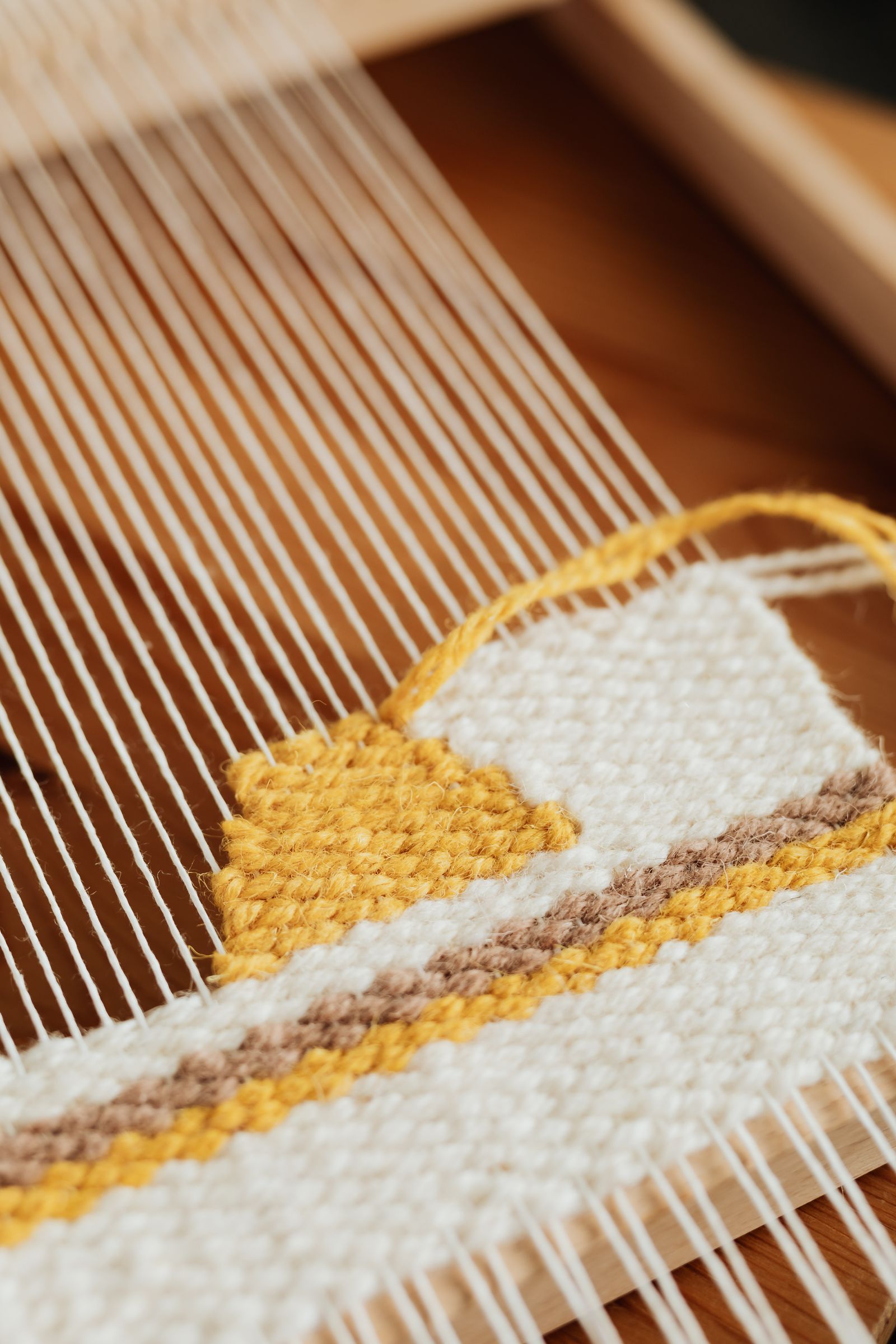 Close-up of cream-white warp threads on a loom with yellow and brown weft interlacing, showing the plain weave structure forming at the fell line